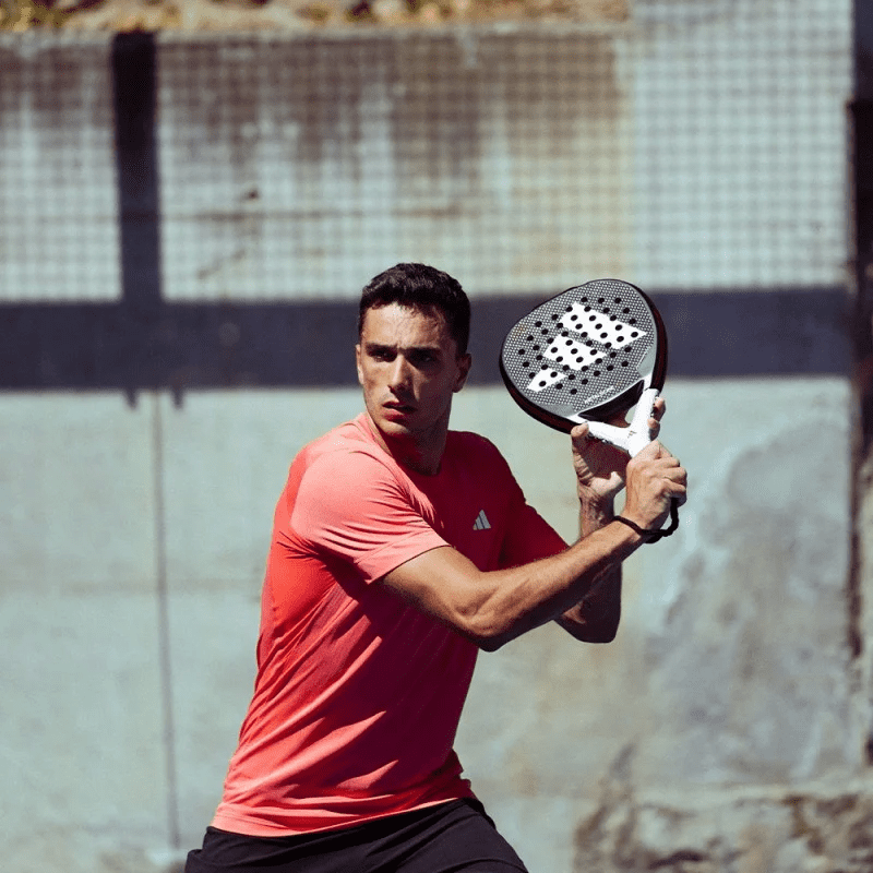 Ein Mann in einem roten Hemd ist bereit, einen Ball auf einem Padelplatz zu schlagen. In der Hand hält er einen Adidas Metalbone HRD+ 2024 Padelschläger. Im Hintergrund sind ein unscharfes Netz und eine Wand zu sehen, was auf eine Umgebung im Freien hindeutet.