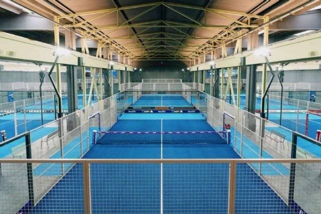 Indoor-Sportanlage mit mehreren parallel angeordneten blauen Padelplätzen, die mit Metallzäunen umschlossen sind. Von oben beleuchtete Leuchtstoffröhren und freiliegende Metallträger säumen die Decke. Besucher fragen oft nach den „Kosten für den Bau eines Padelplatzes“.