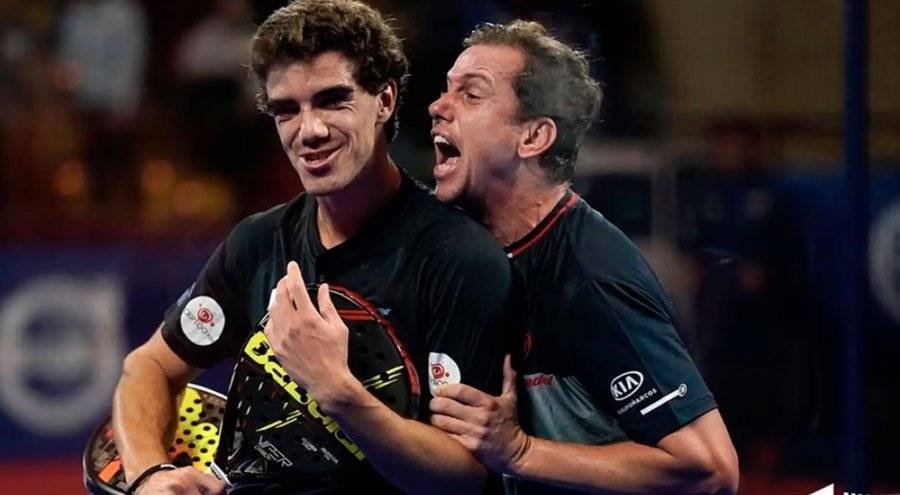 Zwei Männer feiern einen Sieg auf einem Paddle-Tennis-Platz. Ein Mann lächelt und hält einen Schläger in der Hand. Er freut sich, dass sie in der offiziellen Rangliste der Padelspieler der FIP aufgestiegen sind. Der andere steht hinter ihm und jubelt begeistert mit offenem Mund. Beide tragen dunkle Sportshirts.