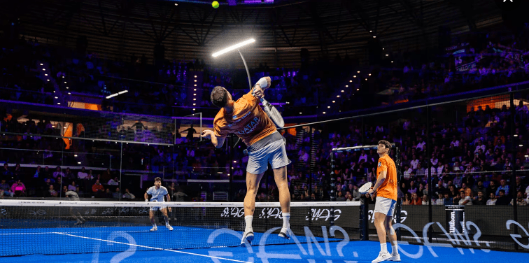 Zwei Tennisspieler in orangefarbenen Hemden und weißen Shorts stehen auf einem Hallenplatz. Ein Spieler springt, um den Ball mit einem Schläger zu schlagen, während der andere zusieht. Der Veranstaltungsort, Teil der Südamerika-Tournee von Premier Padel, ist unter leuchtend violettem Licht voll mit Zuschauern.