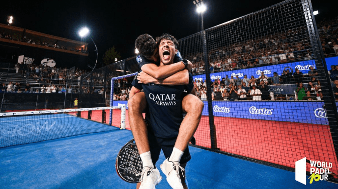 Zwei Padel-Spieler feiern ihren Sieg auf einem Platz, einer springt dem anderen in die Arme. Sie tragen dunkle Uniformen mit einem Sponsorenlogo, während die Zuschauer unter Stadionlichtern jubeln. In der Ecke ist „World Padel Tour“ zu sehen, was ihr Debüt bei der Südamerika-Tournee von Premier Padel markiert.