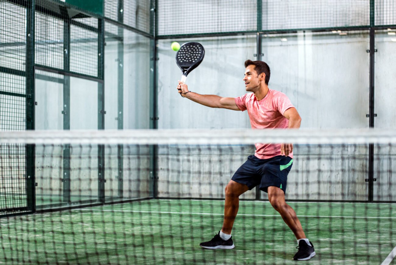 Ein Mann in einem rosa Hemd und dunklen Shorts spielt Padel auf einem grünen Platz und bereitet sich darauf vor, mit seinem schwarzen Schläger einen Ball zu schlagen. Die Szene folgt den fünf Padel-Regeln und spielt sich in einem umzäunten Bereich ab, in dessen Vordergrund sich ein Metallzaun und ein Netz befinden.