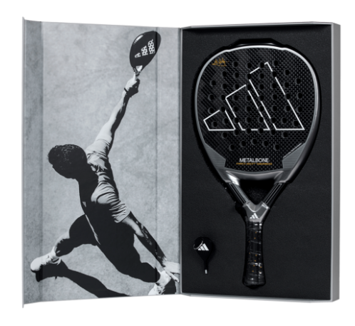 Ein eleganter, schwarzer Adidas Metalbone Padelschläger steht elegant in einer offenen Schachtel neben einer Graustufen-Actionaufnahme von Marta Ortega, die ihren Schläger schwingt. Der Adidas LTD Padelschläger zeigt ein geometrisches Design mit dem ikonischen Logo, das prominent auf dem Griff abgebildet ist.