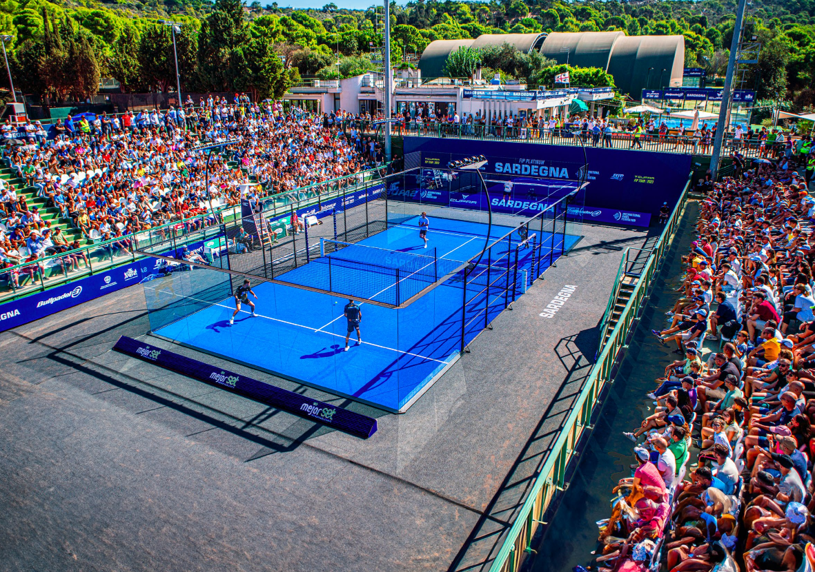 Luftaufnahme eines Padel-Spiels im Freien in einer lebhaften Arena voller Zuschauer. Der Platz ist von bunten Bannern umgeben und zwei Teams spielen bei hellem Tageslicht, was die wachsende Begeisterung für Padel in Deutschland widerspiegelt, mit üppiger grüner Umgebung im Hintergrund.